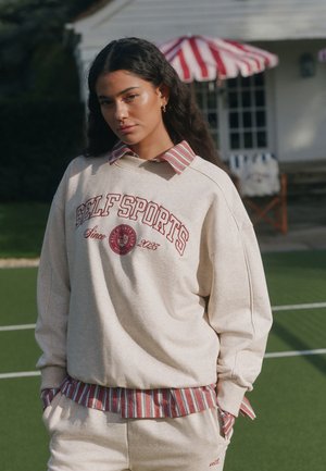 Junge Person mit langen dunklen Haaren, die einen cremefarbenen Sweatshirt mit rotem Text und ein gestreiftes Hemd trägt, steht auf einem grünen Sportplatz in der Nähe eines Hauses.