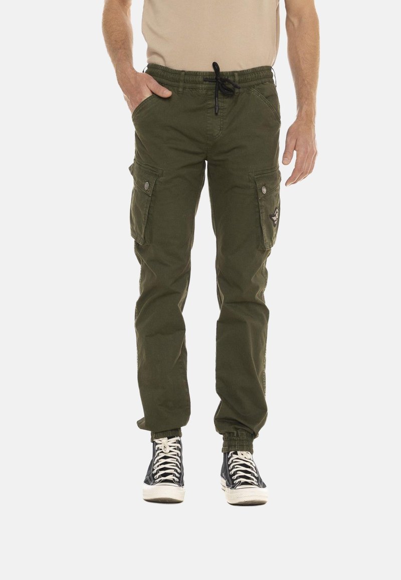 Homme portant un pantalon cargo vert olive à taille coulissée et des baskets montantes noires, debout avec une main dans la poche.