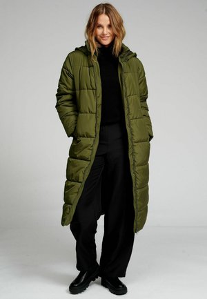 Kvinde stående i en lang olivengrøn dunfrakke, sort højhalset trøje, sorte bukser og sorte støvler mod en ensfarvet grå baggrund.