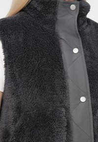 Femme portant une veste polaire texturée gris foncé avec des boutons pression sur une chemise blanche côtelée, montrant le haut du torse et la zone du col.