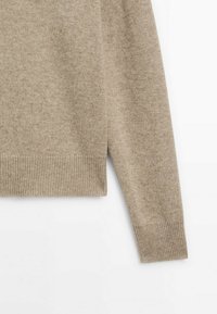 Manche et partie basse du torse d'un pull tricot beige avec poignets et ourlet côtelés sur fond blanc.