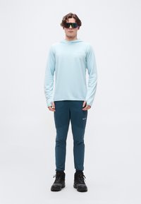 Άνδρας φοράει ανοιχτό μπλε μακρυμάνικο πουκάμισο με κουκούλα, σκούρο μπλε παντελόνι, μαύρα παπούτσια πεζοπορίας και ανακλαστικά αθλητικά γυαλιά ηλίου, στέκεται σε λευκό φόντο.