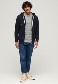 Marineblå zip-hoodie med cremefarvede accenter over en grå t-shirt, parret med blå jeans og beige høj-top sneakers. Krøllet hår og overskæg.