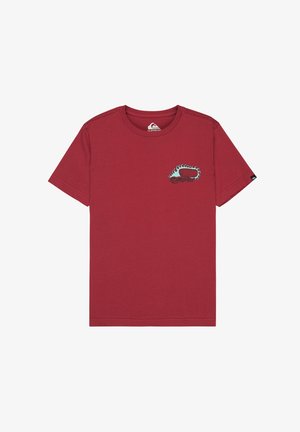 Młoda osoba z kręconymi włosami, ubrana w czerwony t-shirt z nadrukiem, beżowe szorty, białe skarpetki i czarne trampki, stojąca na białym tle.