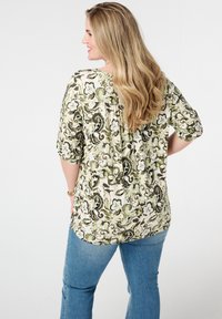 Bloes met bloemenprint in groen en crème tinten, zachte textuur, korte mouwen en een geraffineerde achterkant. Draagt met blauwe jeans.