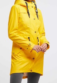 Gul vattentät parka med huva, framknappar, dragsko i midjan och två sidofickor. Har svarta repdetaljer och muddar.
