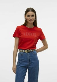 T-shirt en coton rouge avec "ADIOS" imprimé dans une couleur contrastante. Porté avec un jean bleu taille haute et une ceinture rayée, présentant une coupe décontractée.