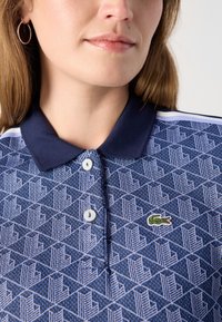 Mėlyna ir tamsiai mėlyna polo marškinėlių su apykakle, pasižyminčių geometriniu raštu, dviem baltomis knopėmis ir mažuoju žaliu krokodilu logotipu.
