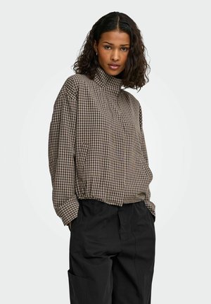 Femme aux cheveux bouclés et foncés portant une veste marron à carreaux à col montant et un pantalon noir ample, debout les mains dans les poches.