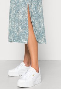 Piernas vistiendo deportivas de plataforma blancas de Puma y una falda corta de un patrón azul claro con una abertura lateral, de pie sobre un suelo gris frente a un fondo blanco.