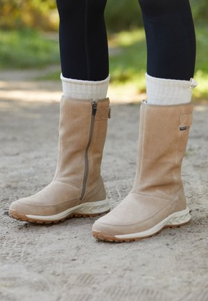 Persona con botas de gamuza beige hasta la mitad de la pantorrilla, calcetines blancos acanalados y leggings negros, de pie en un camino de tierra al aire libre.