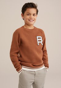 Maglione marrone lavorato a maglia con colletto tondo, polsi e orlo a costine, con un patch bianco e blu con la lettera "R" sul petto. Indossato sopra pantaloni chiari.