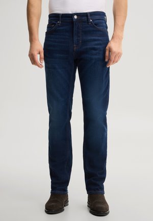 Mann trägt dunkelblaue Slim-Fit-Jeans und dunkelbraune Schuhe, steht mit entspannt an den Seiten hängenden Armen vor einem schlichten hellen Hintergrund.