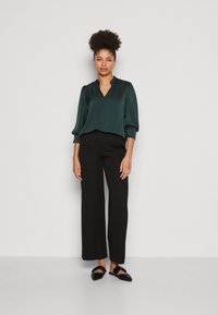 Blouse en satin vert foncé avec un décolleté en V, des manches bouffantes et une coupe décontractée, associée à un pantalon noir à jambes larges et des chaussures noires à enfiler.