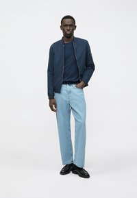 Tmavomodrá bombardérska bunda cez tmavomodré tričko s okrúhlym výstrihom, svetlomodré rifle rovného strihu a čierne topánky. Hladká látka, moderný strih, minimalistický dizajn.