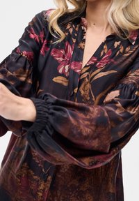 Femme portant un chemisier noir avec des motifs floraux rouges et bruns et des manches volumineuses froncées, montrant le haut du torse et les bras.
