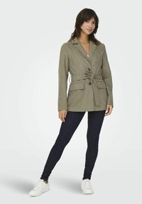 Olijfgroene, ceintuurgsmok met een geknipte kraag, twee voorzakken en knoopsluiting, gecombineerd met donkere jeans en witte sneakers.