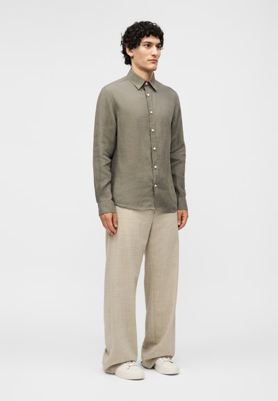Άνδρας όρθιος, φορώντας πουκάμισο ελιάς με κουμπιά, φαρδιά μπεζ παντελόνια και λευκά αθλητικά παπούτσια, ουδέτερο φόντο.