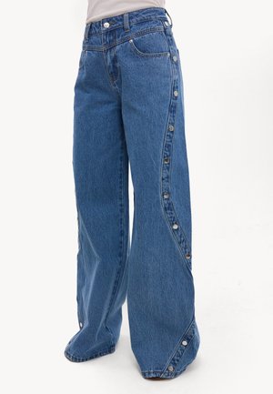 Jean bleu à jambes larges avec des boutons-pression argentés le long des coutures extérieures des jambes et des ourlets, porté avec un haut blanc rentré.