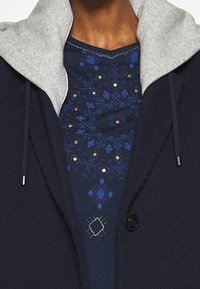 Marineblauer Mantel mit grauer Kapuze; strukturierter Stoff mit Rippenmuster. Darunter ein blaues Hemd mit geometrischen Mustern und goldenen Akzenten.