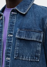 Κοντινό πλάνο τζιν μπουφάν με τσεπάκι στο στήθος με καπάκι, φορεμένο πάνω από μωβ μπλούζα με στρογγυλή λαιμόκοψη.