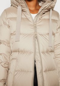 Chaqueta acolchada beige con cierre de cremallera, capucha y tejido plano con textura. Presenta lazos decorativos y un branding minimalista en las correas.