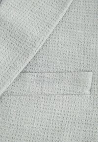 Tejido de color gris claro con textura que presenta un patrón de rejilla sutil, con un borde limpio y un detalle de bolsillo cosido. Material suave y tejido.
