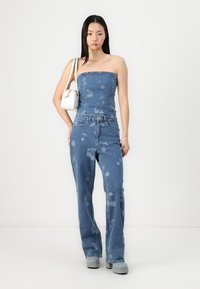 Denim to-delt sett med en strappless topp og flagrende bukser med et lyseblått kirsebærmønster, kombinert med en hvit veske og grå sko.
