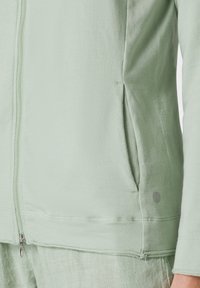 Chaqueta con cremallera de color verde claro, hecha de un tejido suave, con bolsillos laterales, costuras visibles y un pequeño logo cerca del dobladillo.