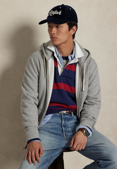 Junger Mann mit schwarzer Kappe, grauem Zip-Hoodie, rot-navy gestreiftem Rugbyshirt, hellblauen Jeans und Armbanduhr, sitzend auf einem Hocker.