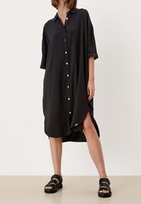 Czarna sukienka koszulowa z krótkimi rękawami, pionowymi paskami, bocznymi rozcięciami i zaokrąglonym dołem. Noszona z czarnymi masywnymi sandałami.