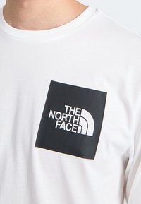 Weißes Baumwoll-T-Shirt mit einem quadratischen schwarzen Logo mit "THE NORTH FACE" und einem geschwungenen weißen Akzent. Runder Ausschnitt, kurze Ärmel.