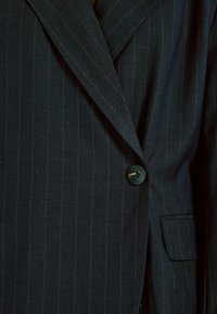 Laivasto sininen pinstripe bleiseri, valmistettu kankaasta, jossa on viillotettu kaulus, yksittäinen nappisulku ja sivutasku läpällä.