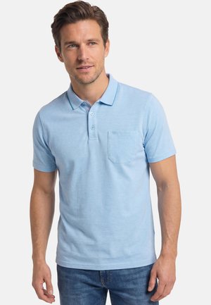 Mann trägt hellblaues kurzärmeliges Poloshirt mit Brusttasche, steht vor einem schlichten weißen Hintergrund und schaut leicht nach rechts.