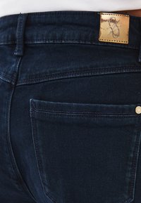 Ciemnoniebieskie dżinsy z denimu z złotą skórzaną łatą na pasie, kontrastowym przeszyciem oraz małą tylną kieszenią z nitką.