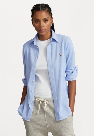 Chemise bleu clair à boutons en tissu doux, avec des manches longues retroussées au niveau des coudes et un petit logo brodé sur la poitrine.