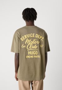 Oliven grøn bomulds t-shirt med stor gul tekst på ryggen, der står "SERVICE DEPT. MOTOR CLUB EST. ’67 HUGO ENGINE PARTS."
