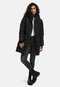 Abrigo puffer negro con capucha, con un patrón acolchado, cremallera frontal y cordones. Combinado con jeans ajustados grises y botas negras de tobillo.