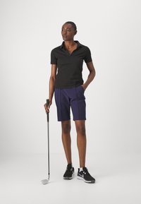 Cămașă polo neagră cu guler și detalii cu nasturi, asortată cu șorturi violet până la genunchi. Modelul ține un club de golf, purtând pantofi sport negri.