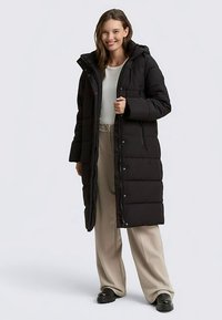 Zwart gewatteerde jas met capuchon, voorzien van een doorgestikt ontwerp, knopen aan de voorkant en steekzakken aan de zijkant, gedragen over lichtgekleurde broek.