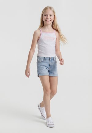 Fată tânără care merge, purtând un top alb fără mâneci cu margine roz, pantaloni scurți din denim bleu-deschis și adidași albi, zâmbind pe un fundal simplu.