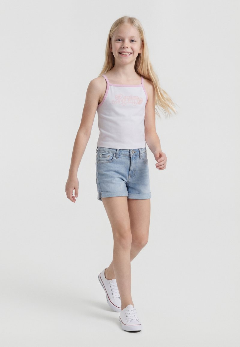 Μικρό κορίτσι περπατάει, φορώντας ένα λευκό αμάνικο τοπ με ροζ λεπτομέρεια, ανοιχτό μπλε τζιν σορτς και λευκά αθλητικά παπούτσια, χαμογελώντας σε μονόχρωμο φόντο.