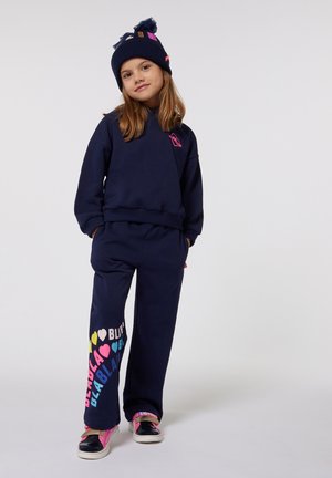Outfit composto da felpa navy e pantaloni con dettagli rosa; i pantaloni presentano un testo grafico colorato "I love BLABLA." Il modello indossa un berretto abbinato.