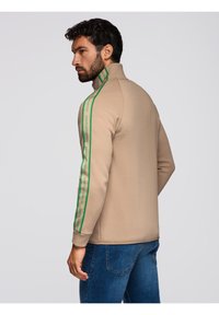 Felpa beige con colletto alto e strisce laterali verdi con testo, realizzata in materiale morbido, abbinata a jeans in denim blu.