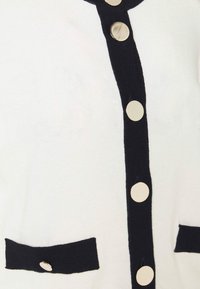 White cardigan with black trim, featuring round metallic buttons and two black pockets. Knit fabric with a smooth texture.