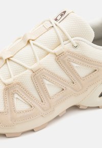 Beige athletic shoe with mesh upper and textured overlays. Features a lace system and triangular cutouts for ventilation and design.