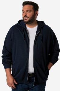 Felpa con zip di colore navy, con cappuccio e polsini a coste, caratterizzata da una superficie liscia e un logo discreto sul petto, indossata sopra una t-shirt bianca.