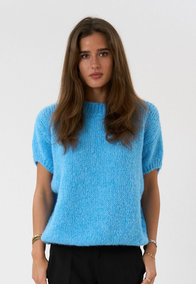 Jeune femme aux longs cheveux bruns portant un pull court à manches courtes et duveteux bleu clair et un pantalon noir, sur un fond gris uni.
