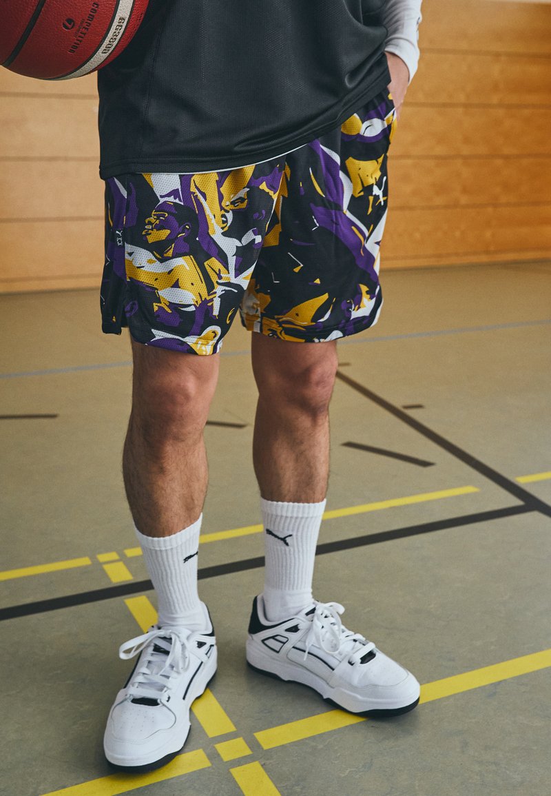 Pantalones cortos de baloncesto en un estampado abstracto de color negro, púrpura y amarillo. Llevados con calcetines blancos y zapatillas deportivas blancas. Ambiente de gimnasio con un balón de baloncesto.