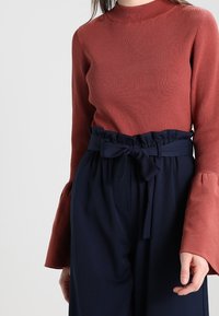 Rostfarbener, gerippter Oberteil mit hohem Kragen und Glockenärmel, kombiniert mit navyblauen, hochtaillierten Hosen mit gebundenem Bund.